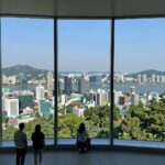 남산타워 전망대에서 바라본 서울 시내와 한강이 한눈에 들어오는 파노라마 뷰, 360도 유리창 너머 보이는 고층 빌딩과 푸른 숲이 어우러져 있다.